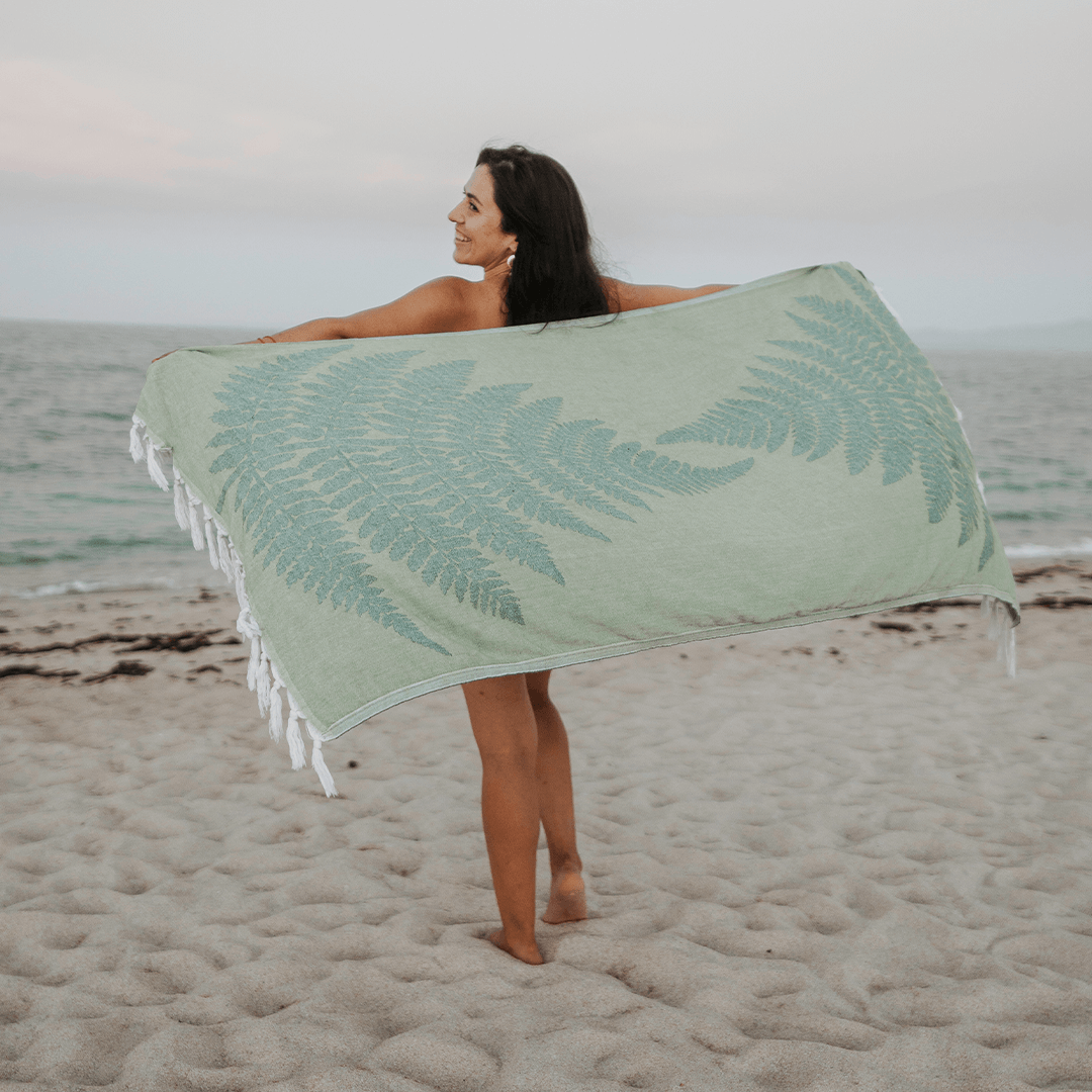A woman holding up a green fern Pomp & Sass towel while walking on the beach.