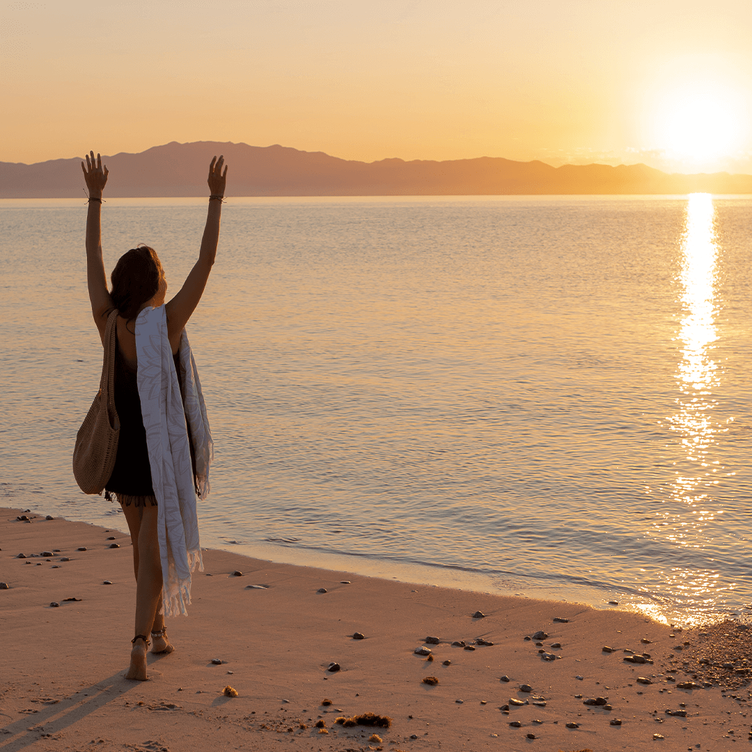 Awoman rasing her hands up while watching the sunset at the beach. She has a white Turkish towel draped over he shoulder.