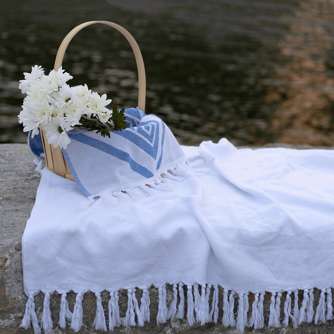 A white Turkish towel placed on a rock beside the lake used as a blanket, on top there is a basket holding a white flower and a blue diamond patterned Turkish towel.
