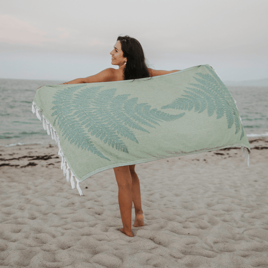 A woman holding up a green fern Pomp & Sass towel while walking on the beach. 