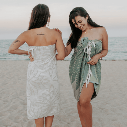 Two women on the beach draped in a green fern Turkish towel and a cream coloured Lotus designed Turkish towel.