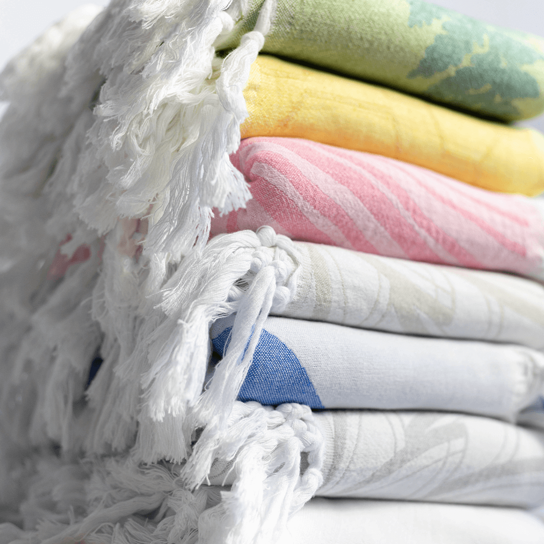 A stack of all the Pomp & Sass towels, green fern, yellow honeycomb, pink striped, cream, blue and white.