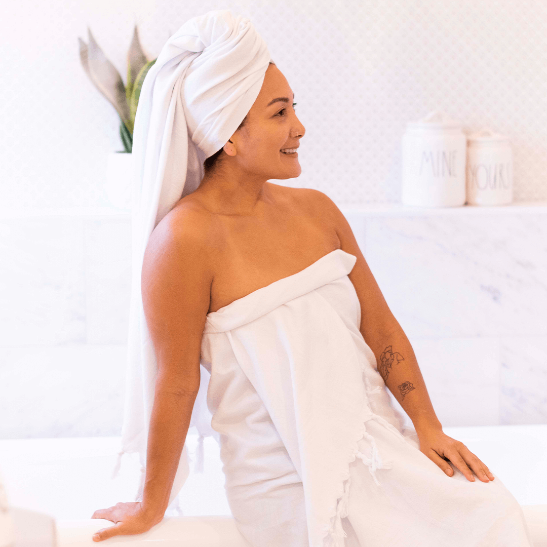 A Latina woman wrapped in a white Turkish towel as well as one on her head, laughing and holding her hand up. 