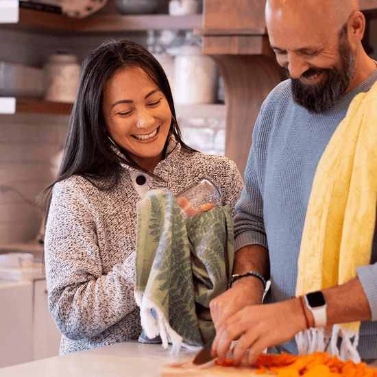 A Latina woman and bearded man cooking and cutting vegetables with green fern and yellow honeycomb Turkish towels draped over their shoulders. One is using the towel to clean off a glass. 