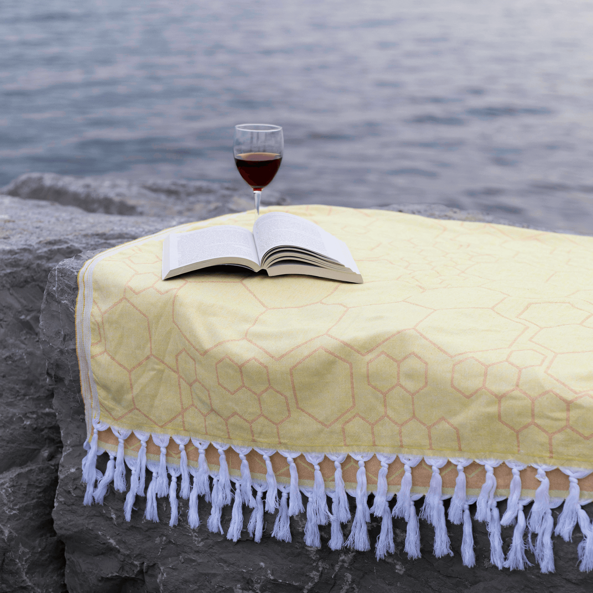 A yellow honeycomb Turkish towel lays on large rocks by the lake. A book and glass of red wine sits with them.