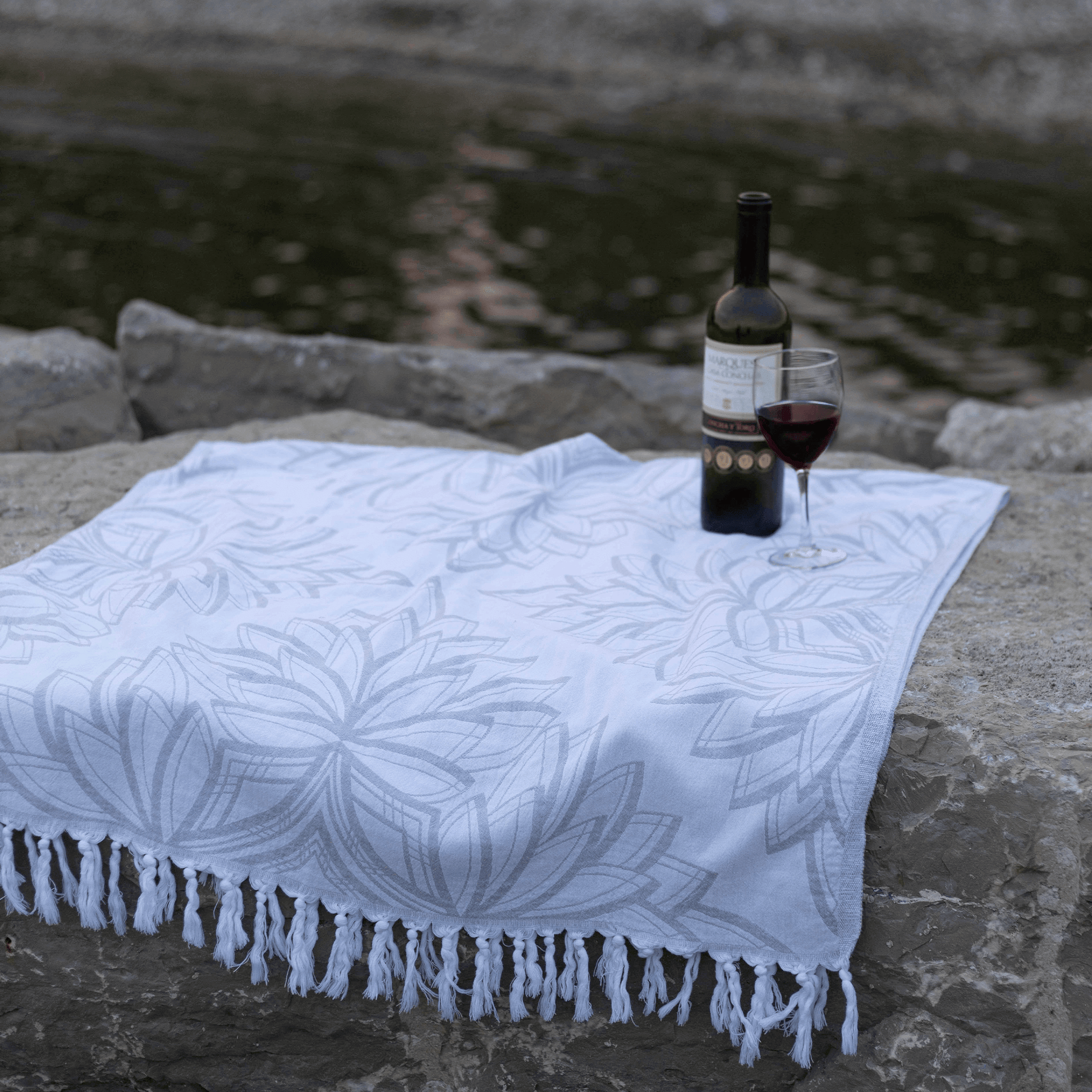 Grey and white Turkish towel on large rocks by a lake with a bottle and glass of red wine