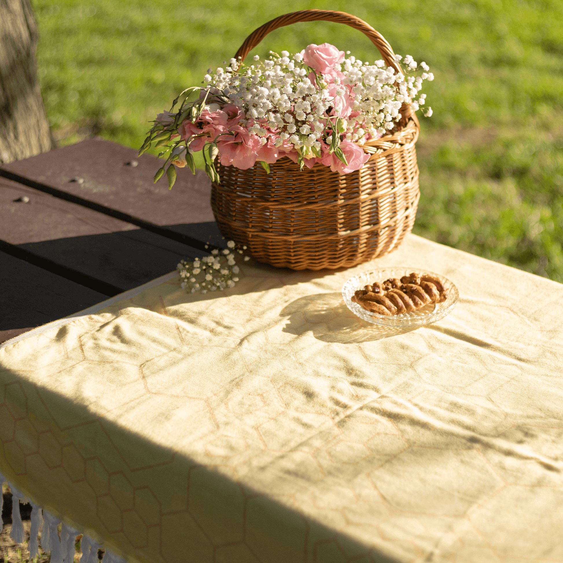 A yellow Turkish towel is used as a picnic table cloth on a summers day in the park. The table also has a basket of flowers and pastry.
