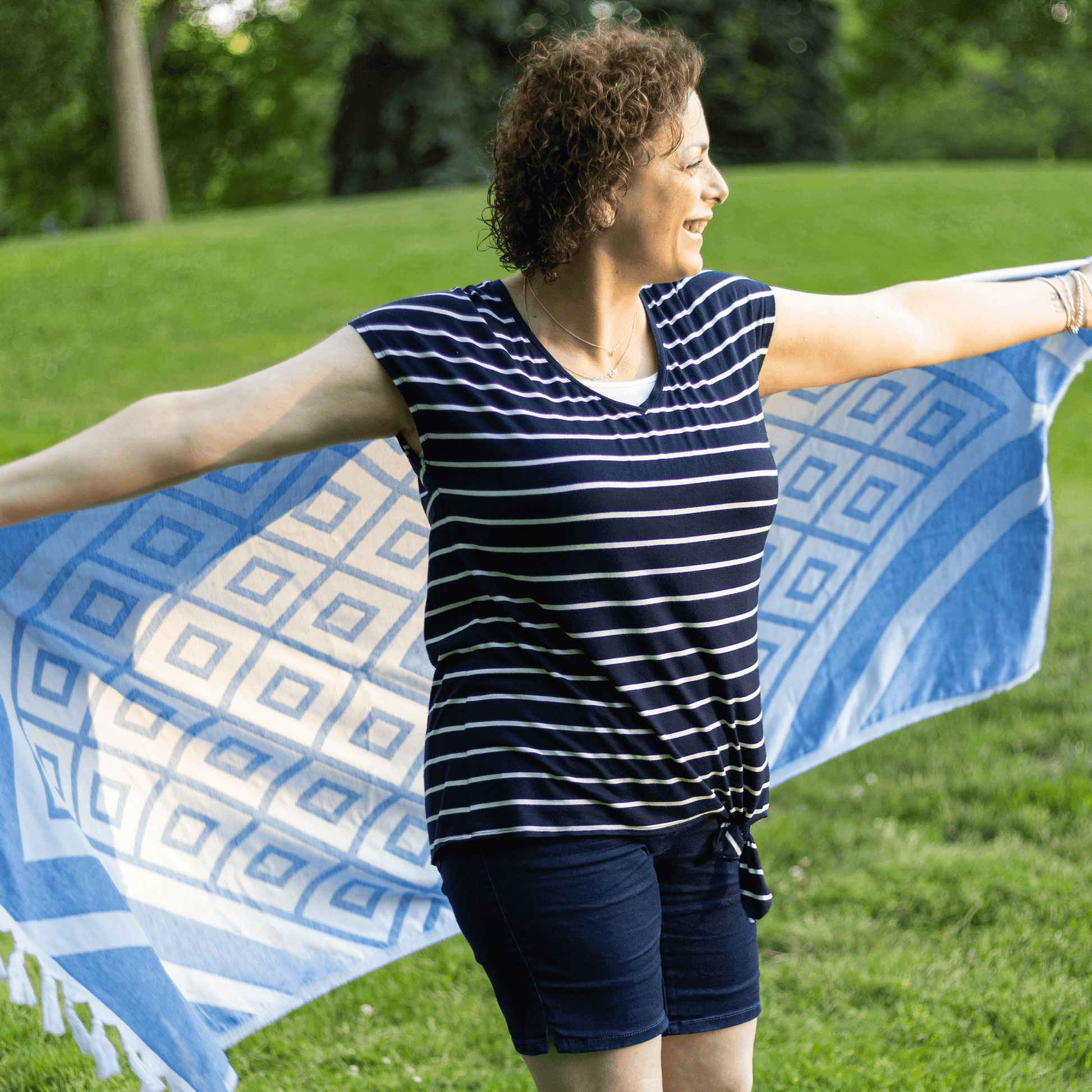 Smiling woman holds out a blue and white Turkish towel outside by green grass