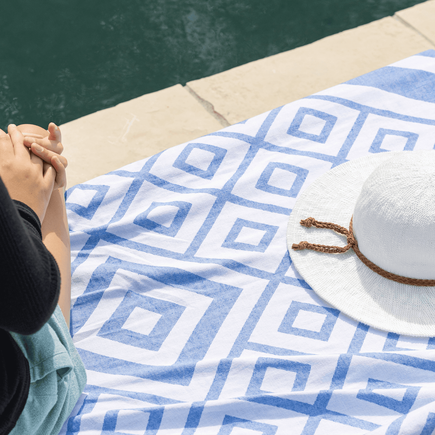 Person sits on a blue and white Turkish towel poolside with a white hat next to them